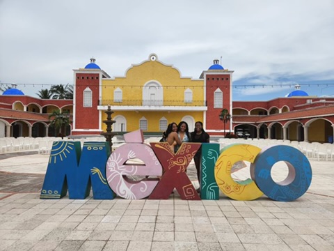 Two women smile and pose in front of the colorful "Mexico" sign