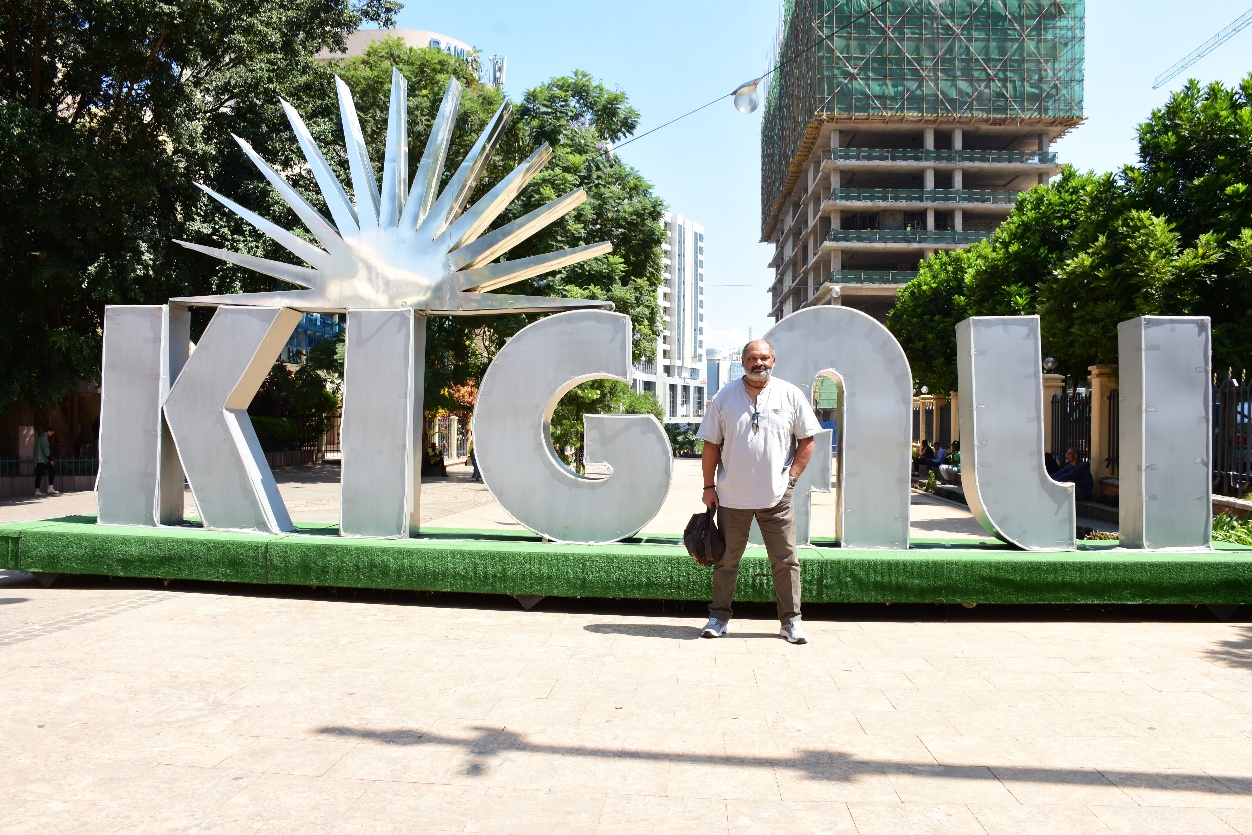 A man stands in front of a large sign displaying the word "Kigali