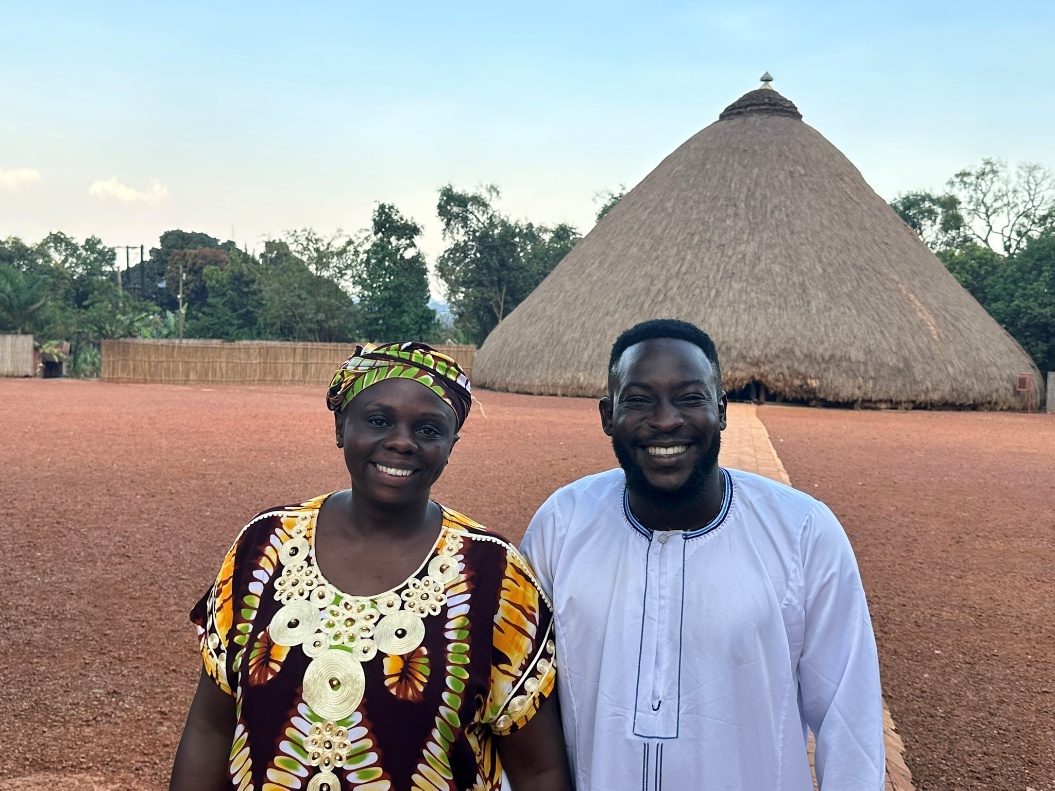 A man and woman stand together in front of a rustic hut
